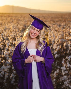 Graduate in purple cap and gown poses in front of a cotton field at sunset during senior portrait session.
