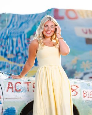 A woman in a yellow sundress poses cheerfully in front of a colorful graffiti wall on a sunny day. Salvation Mountain portrait photography.