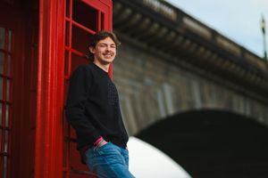 A person in jeans and a dark jacket stands in front of red telephone booths in an urban setting. Senior portraits by Dusty Wooddell