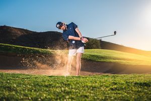 Golfer hitting from sand bunker creating dramatic dust cloud at sunset on golf course. Portrait photography by Dusty Wooddell.