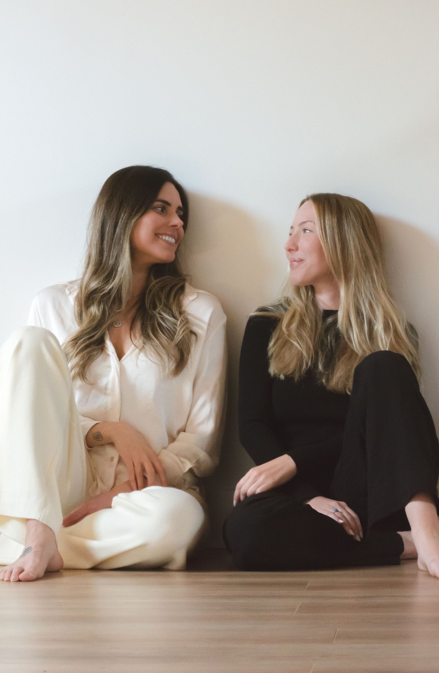 Two women sitting on floor against white wall sharing a candid moment and smiling.