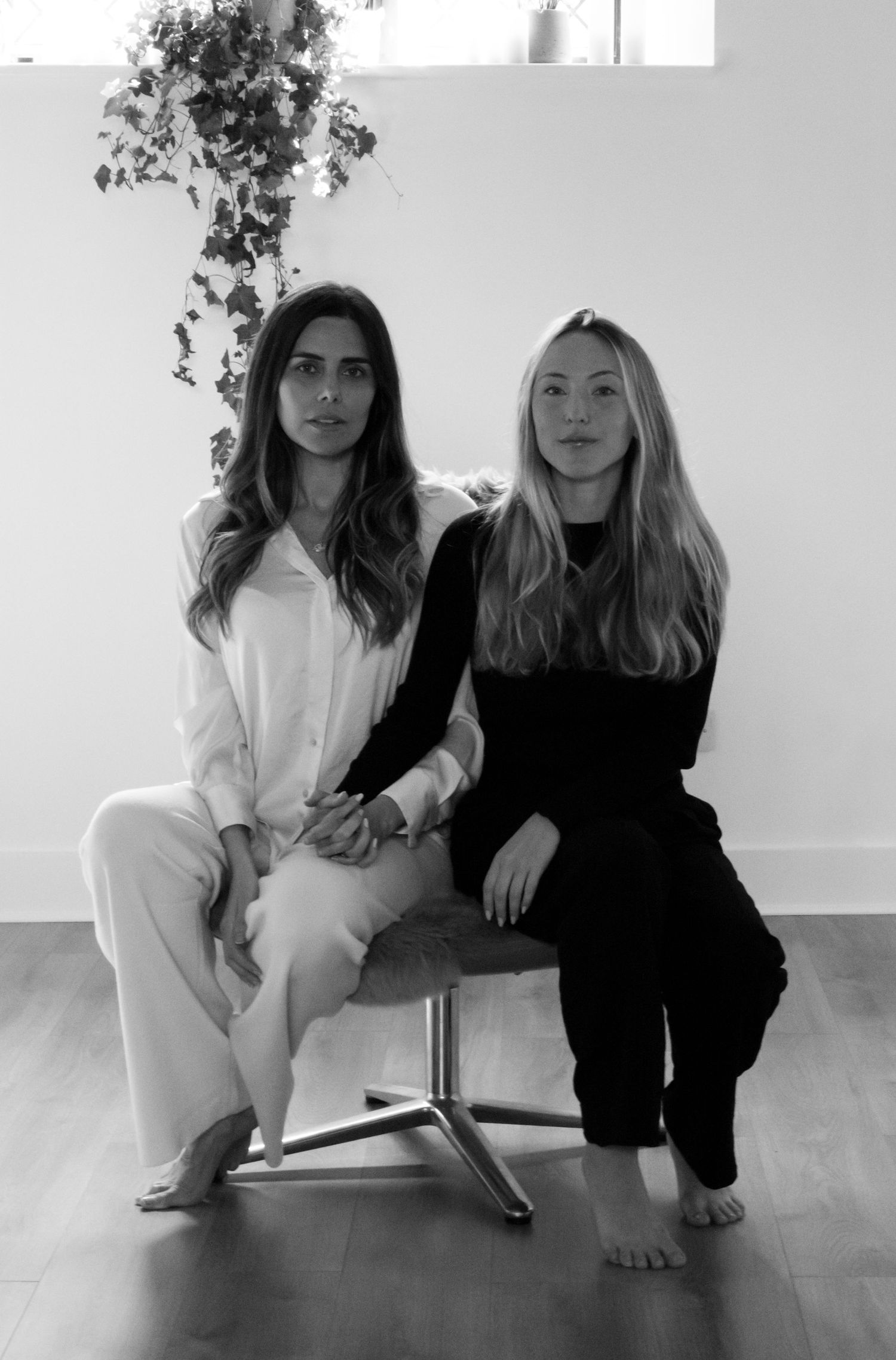Two models in contrasting black and white outfits pose together on a hardwood floor in a minimalist setting.