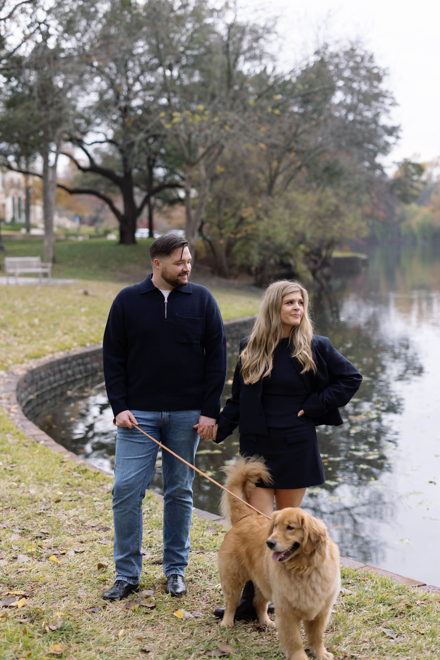 A couple takes their golden retriever for a walk along a scenic pond on an autumn afternoon.