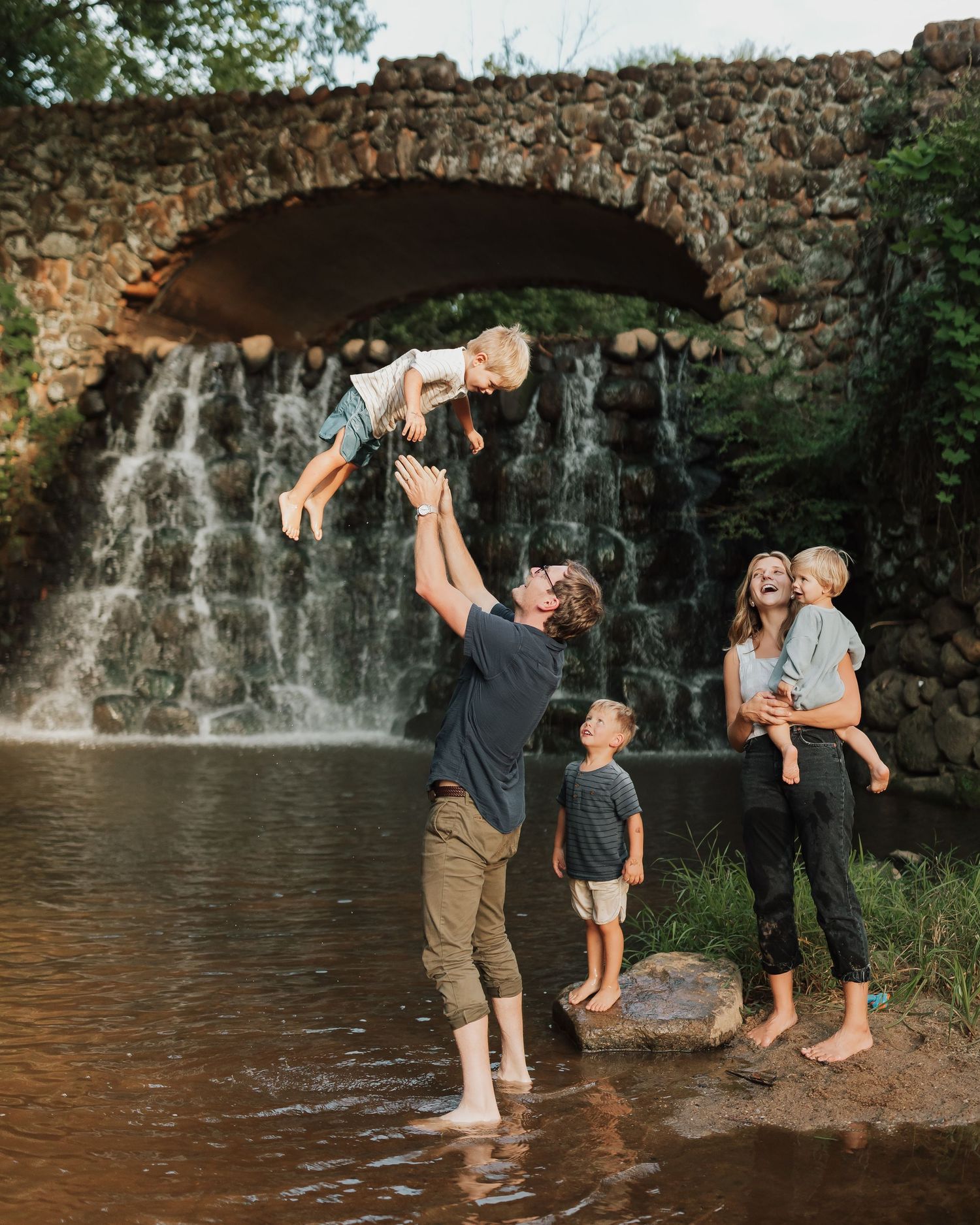 Family enjoys summer day at a scenic stone bridge waterfall, splashing and playing in the water as water cascades behind them.