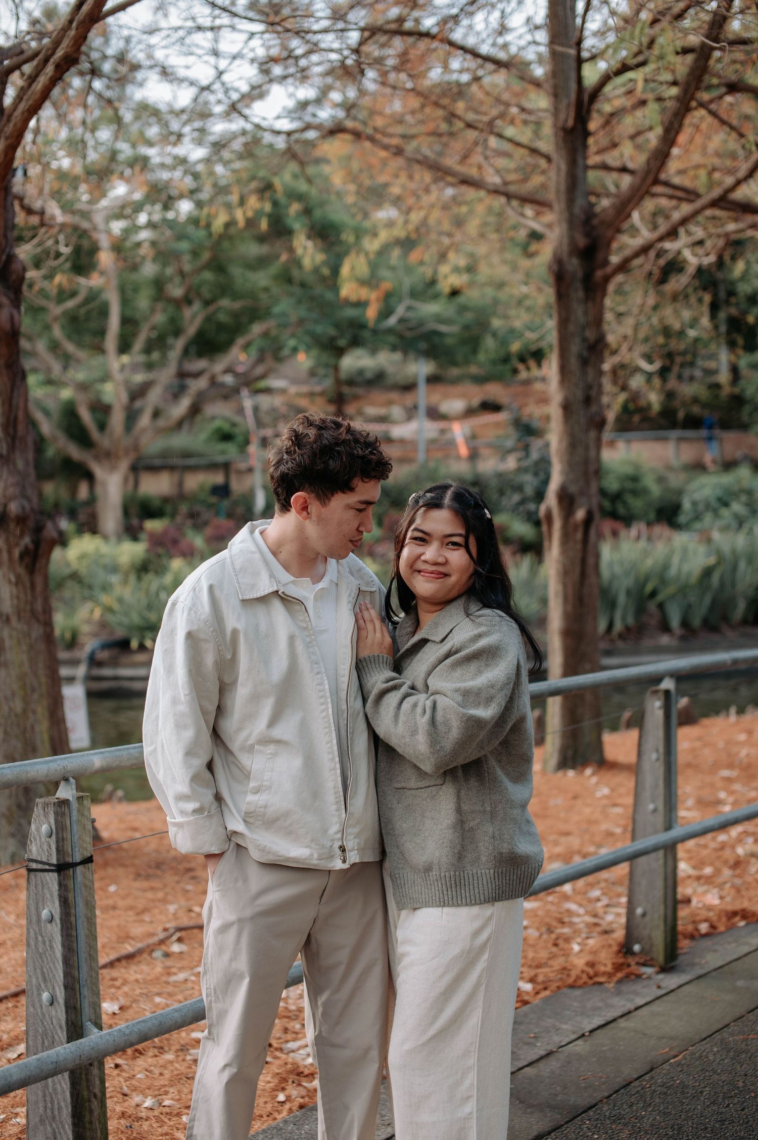 A couple in neutral-colored outfits embraces on a garden pathway during autumn.
