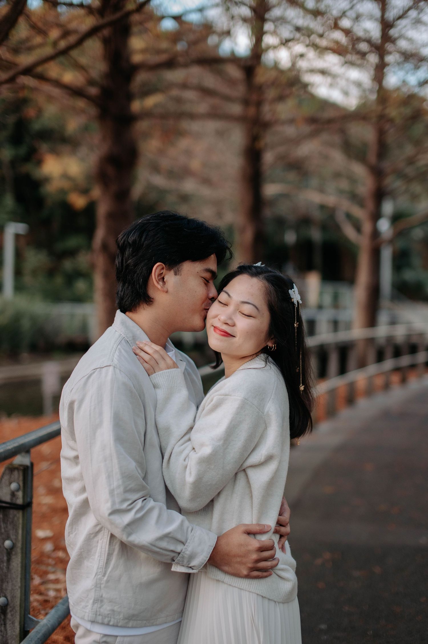A romantic couple in matching white outfits share an intimate moment on a tree-lined path.