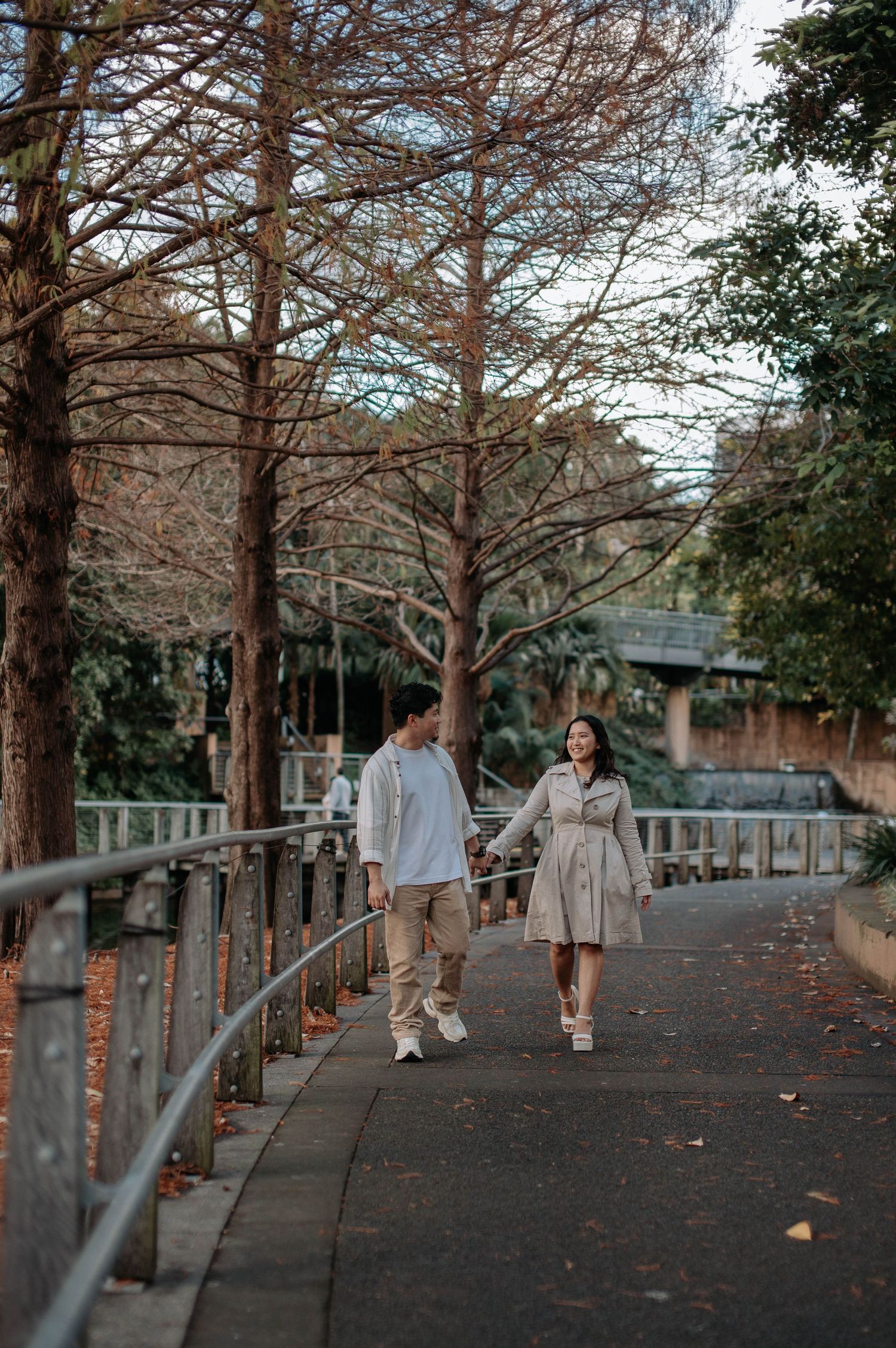 A couple takes a romantic stroll along a tree-lined pathway wearing coordinated light-colored outfits.