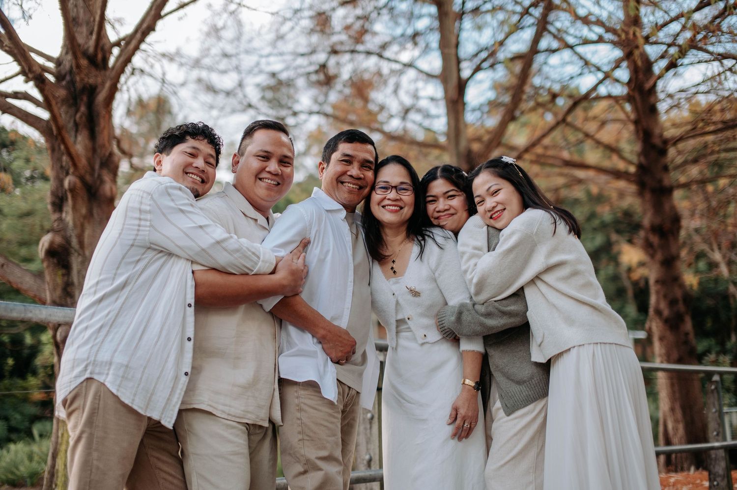 A group in coordinated white and beige outfits poses together on a walkway surrounded by trees.