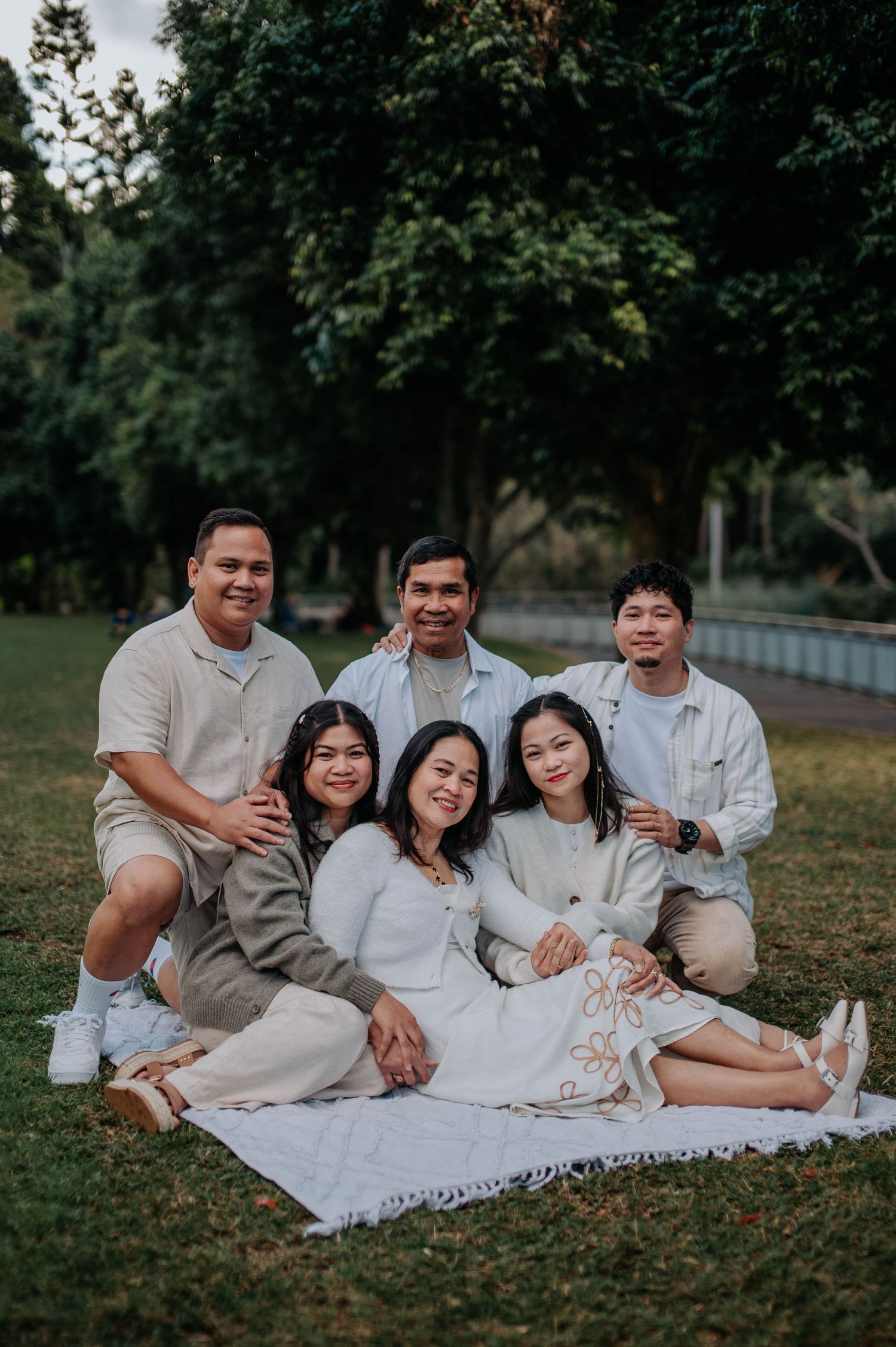 A family group sits together for a casual outdoor portrait on a white blanket in a park setting.