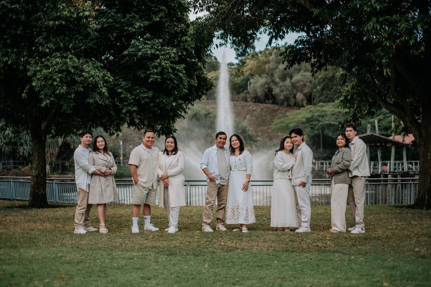 Group in coordinated white and beige attire stand together for portrait in lush park setting.