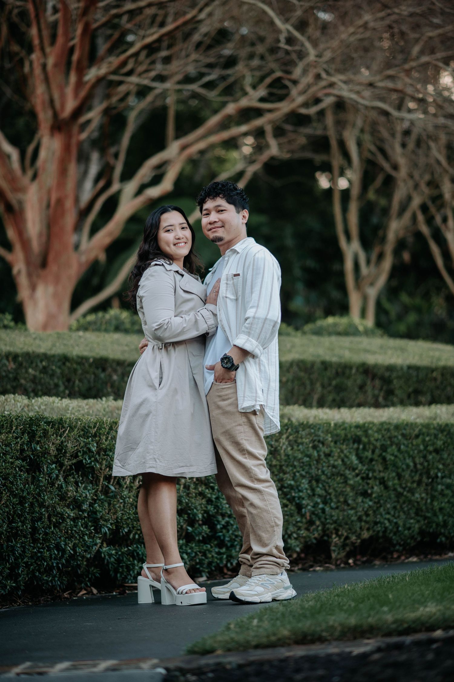A couple in light neutral outfits poses playfully together against a garden backdrop with hedges and trees.