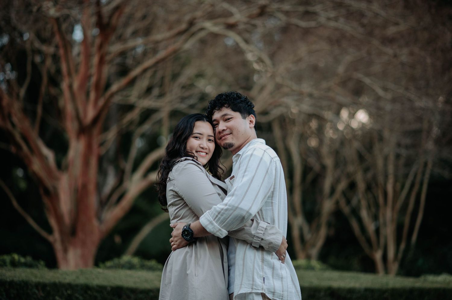 A couple embraces while sharing a tender moment in a park with trees in the background.