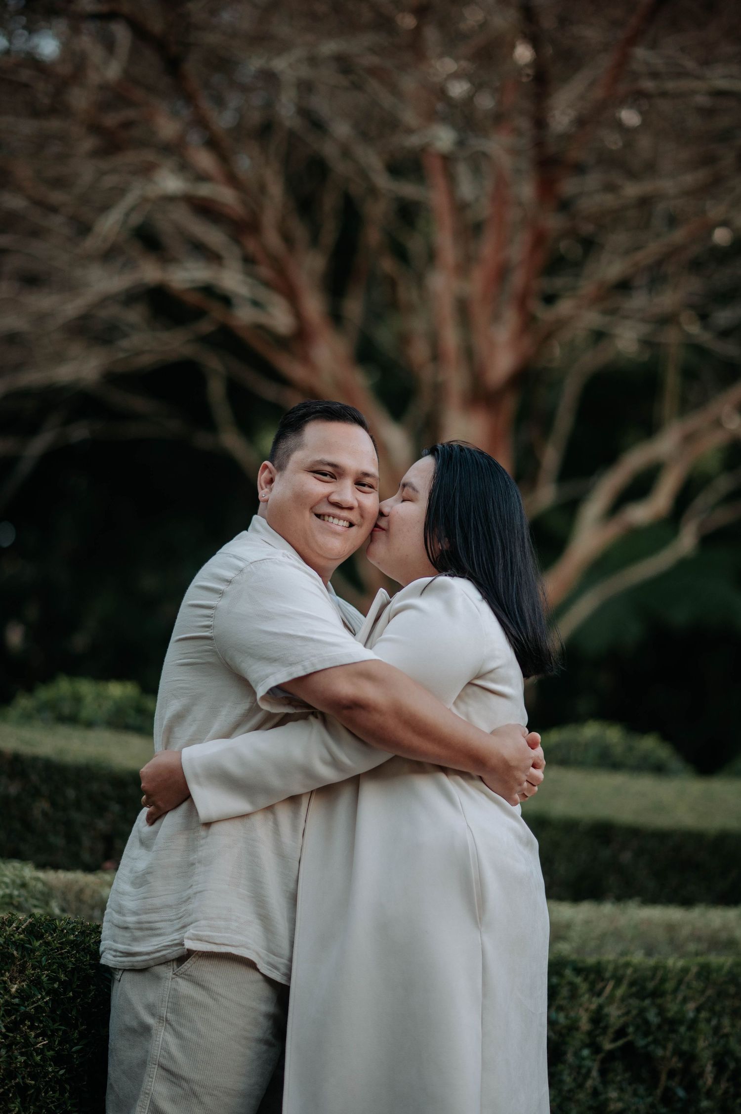 Couple embraces warmly while wearing matching white outfits in front of tree branches.