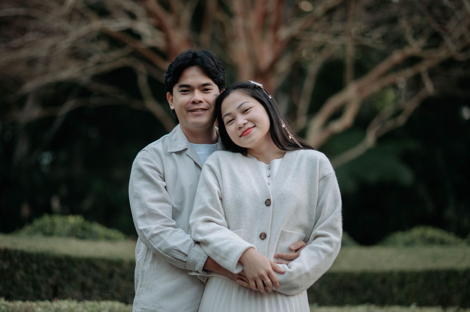 A tender romantic photoshoot in a garden setting shows two people embracing in matching beige outfits.
