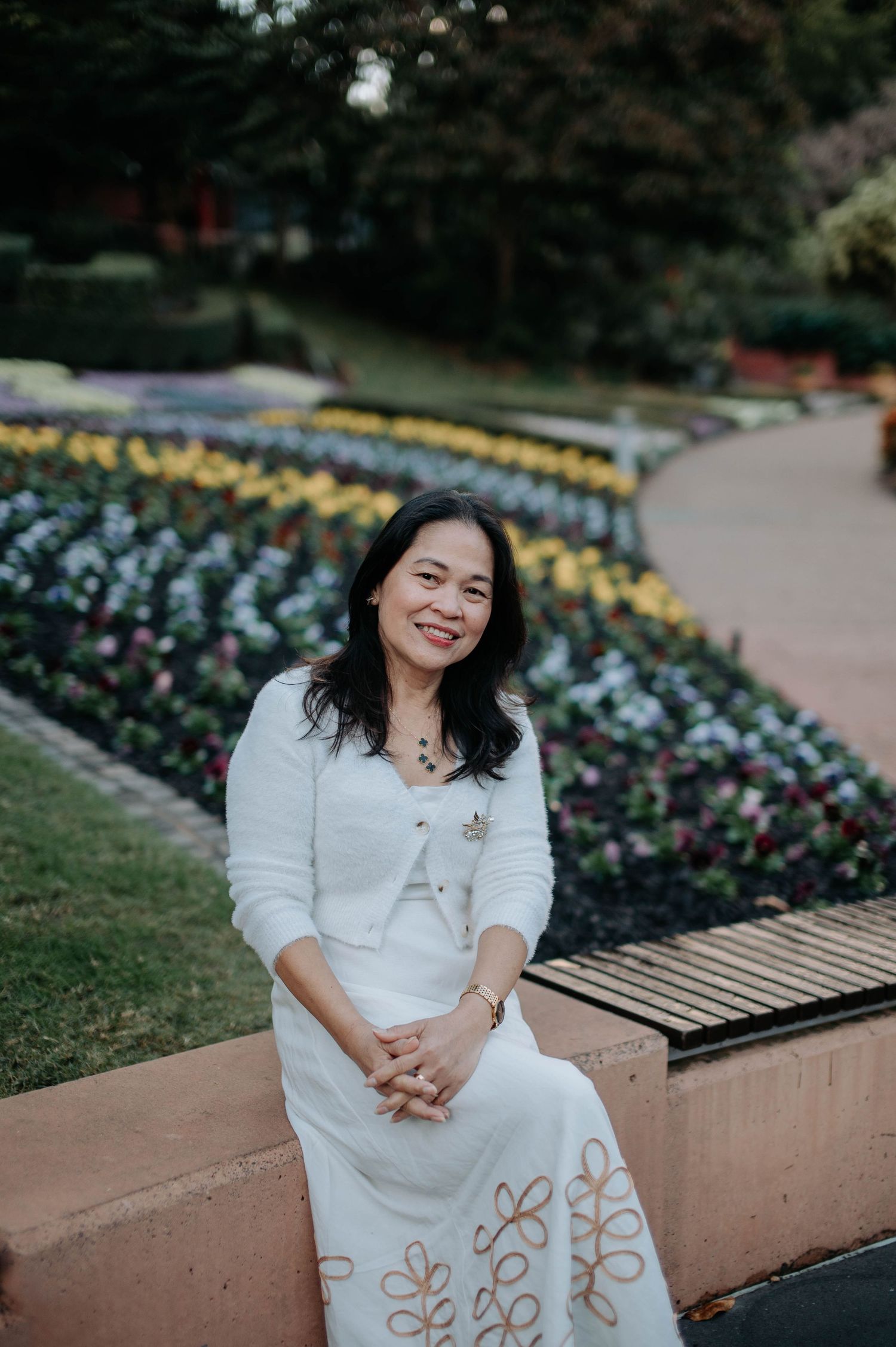 Person in white dress sits on garden wall near colorful flower beds at dusk.