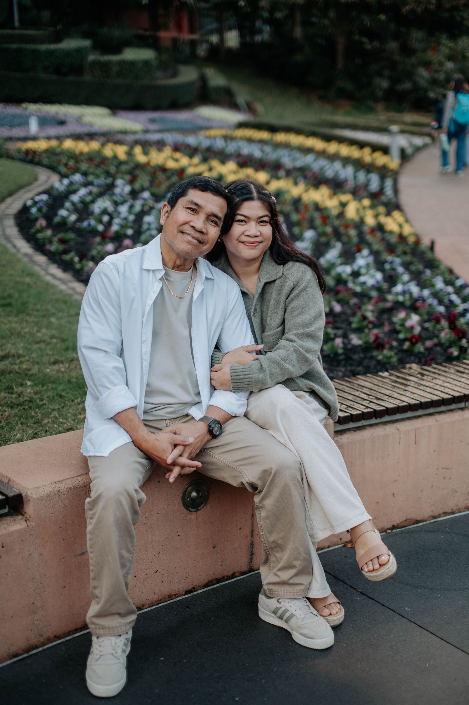 A couple sits together on a garden wall with colorful flower beds in the background.
