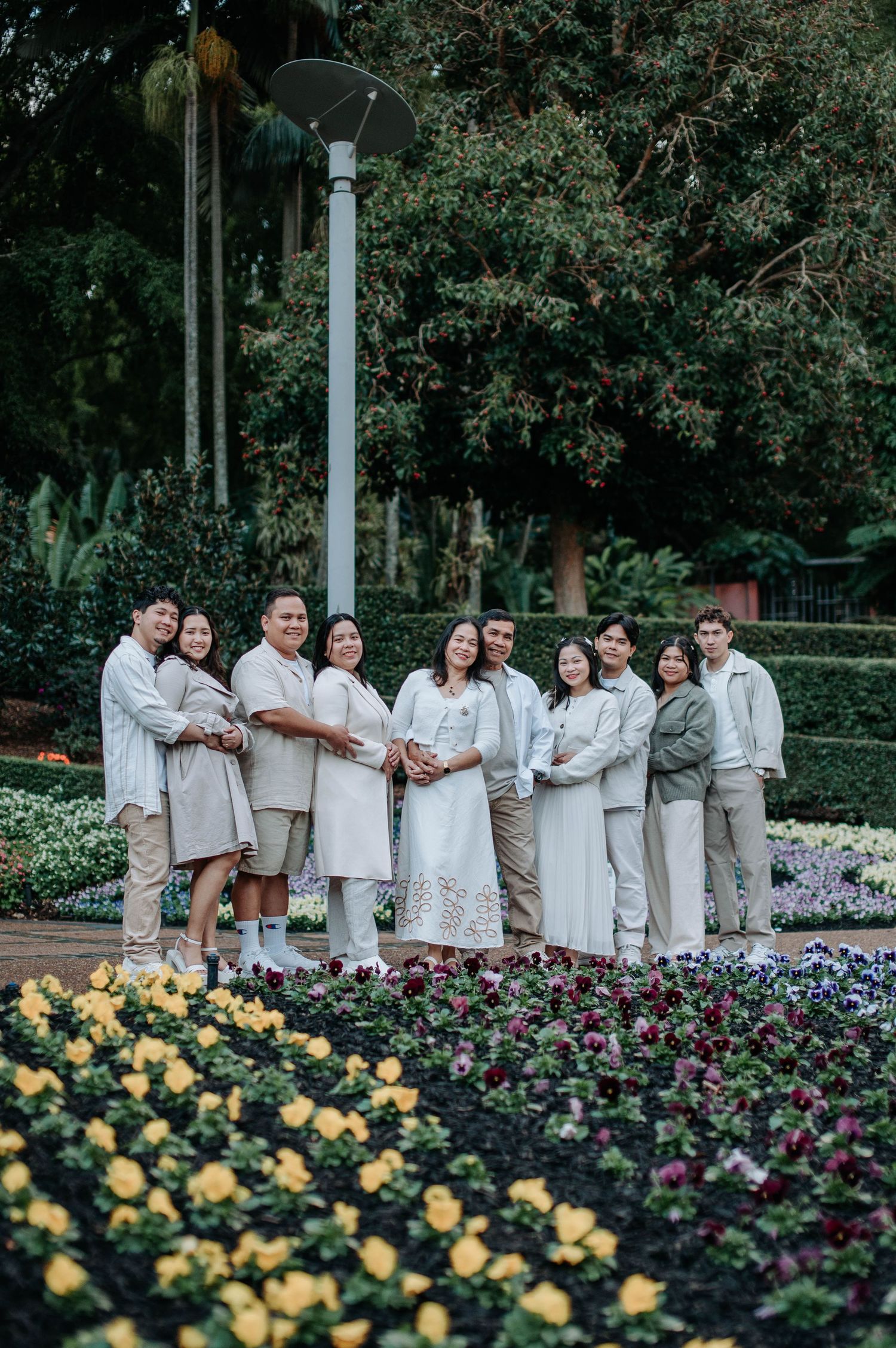 A group photo of people wearing coordinated white and beige outfits standing by a colorful flower garden.