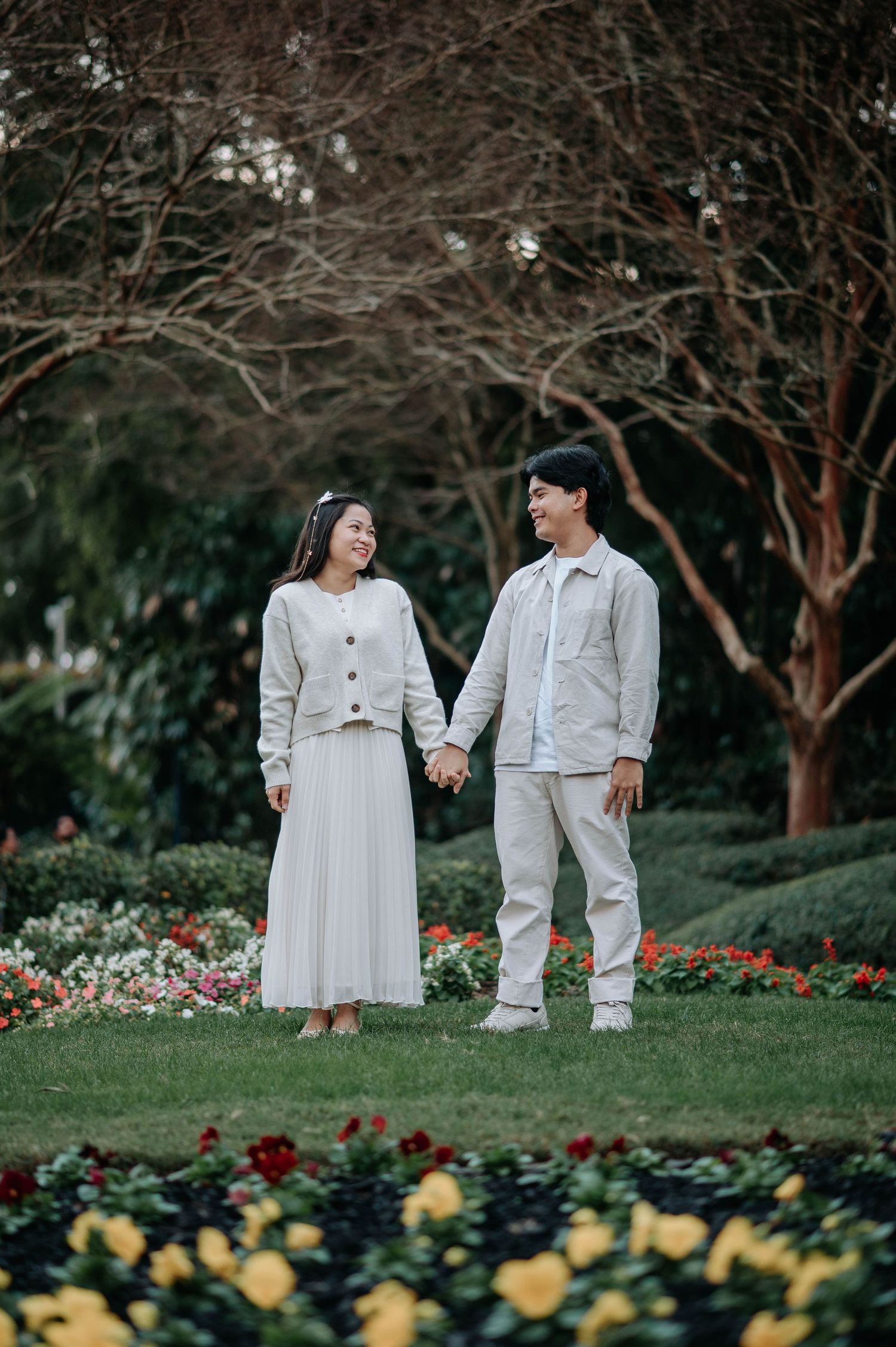 A couple in light-colored outfits holds hands while standing in a garden with blooming yellow flowers.