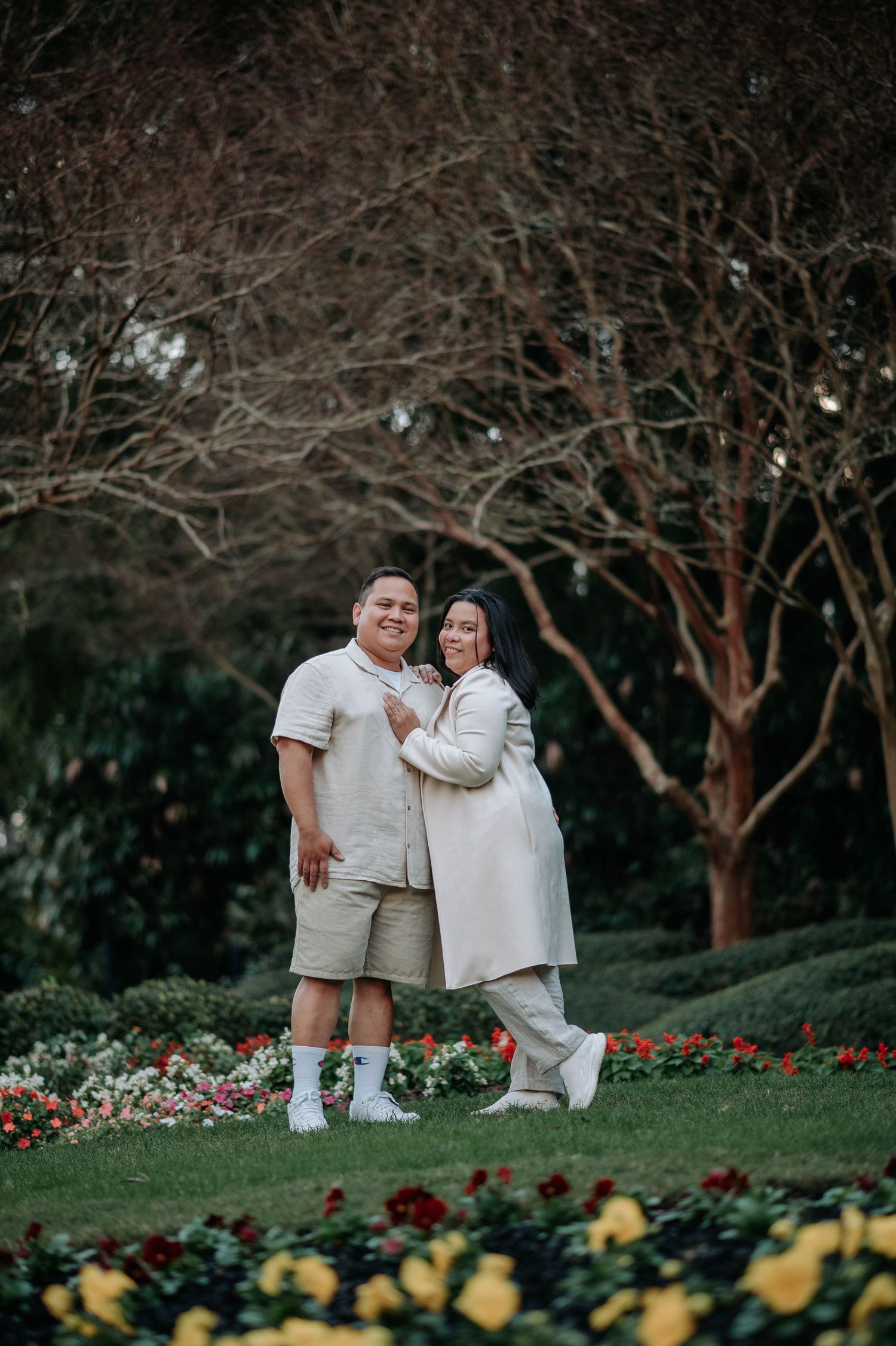 Couple in coordinated white outfits pose together on grassy slope with yellow flowers blooming below.