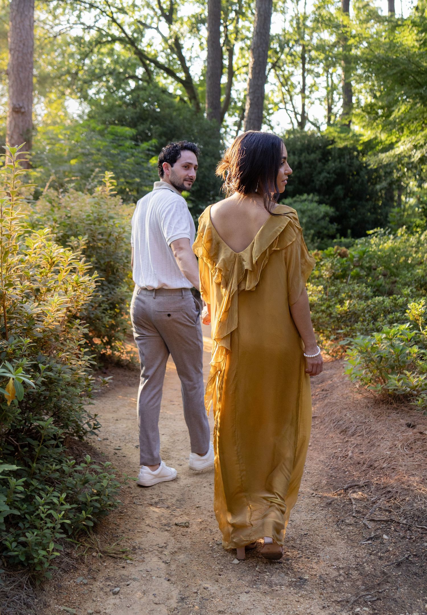Couple walking on a forest trail with golden sunlight filtering through trees.