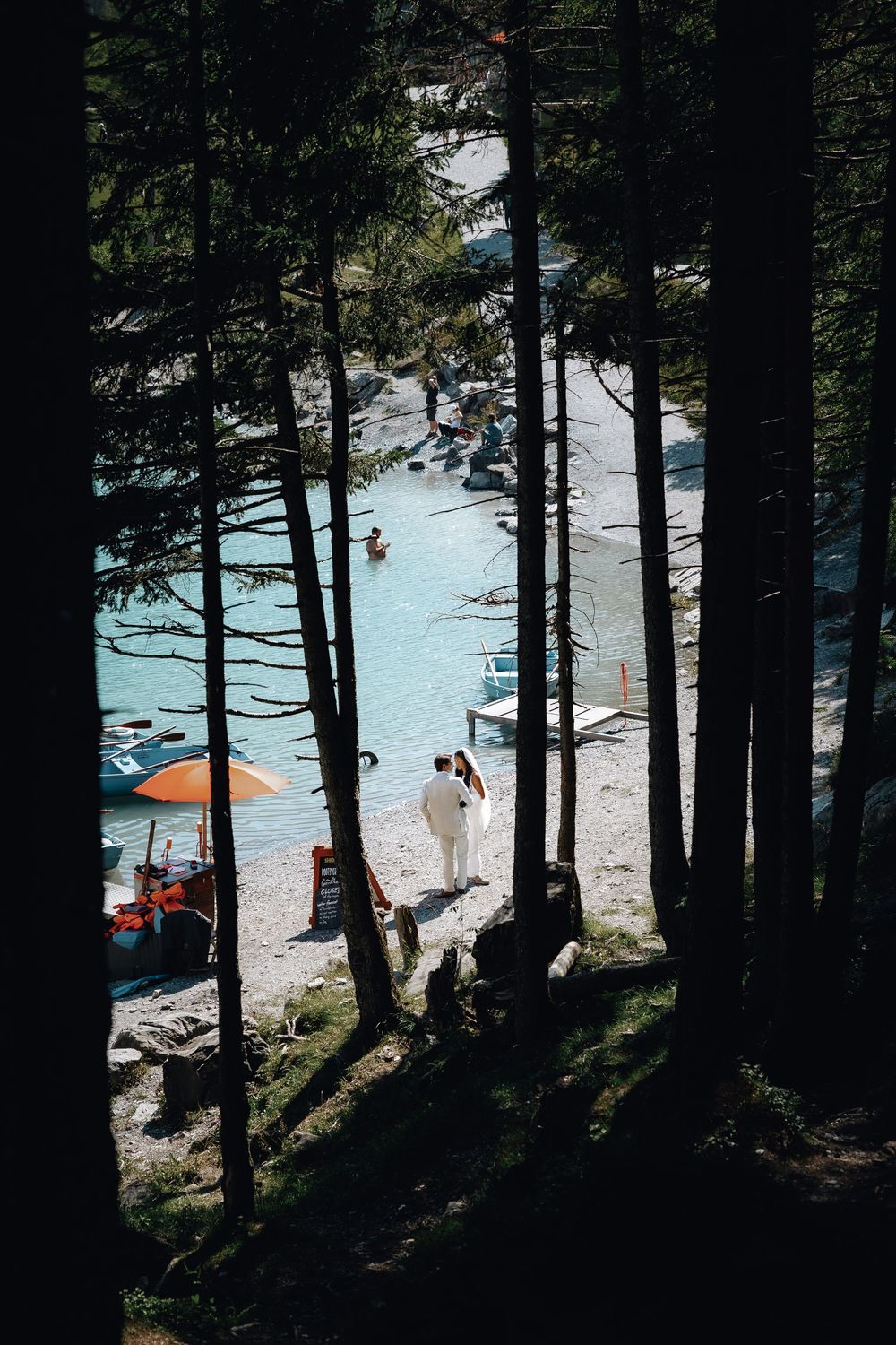 Pine trees frame a lakeside beach with umbrellas and beachgoers visible through the forest.