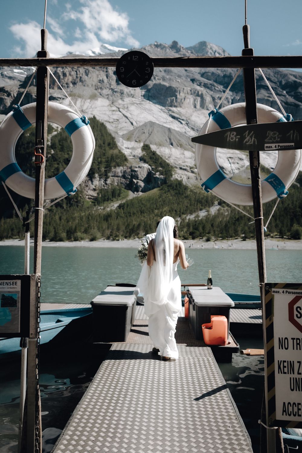 Bride in flowing white dress walks along a boat dock with mountain lake and snow-capped peaks in the background.