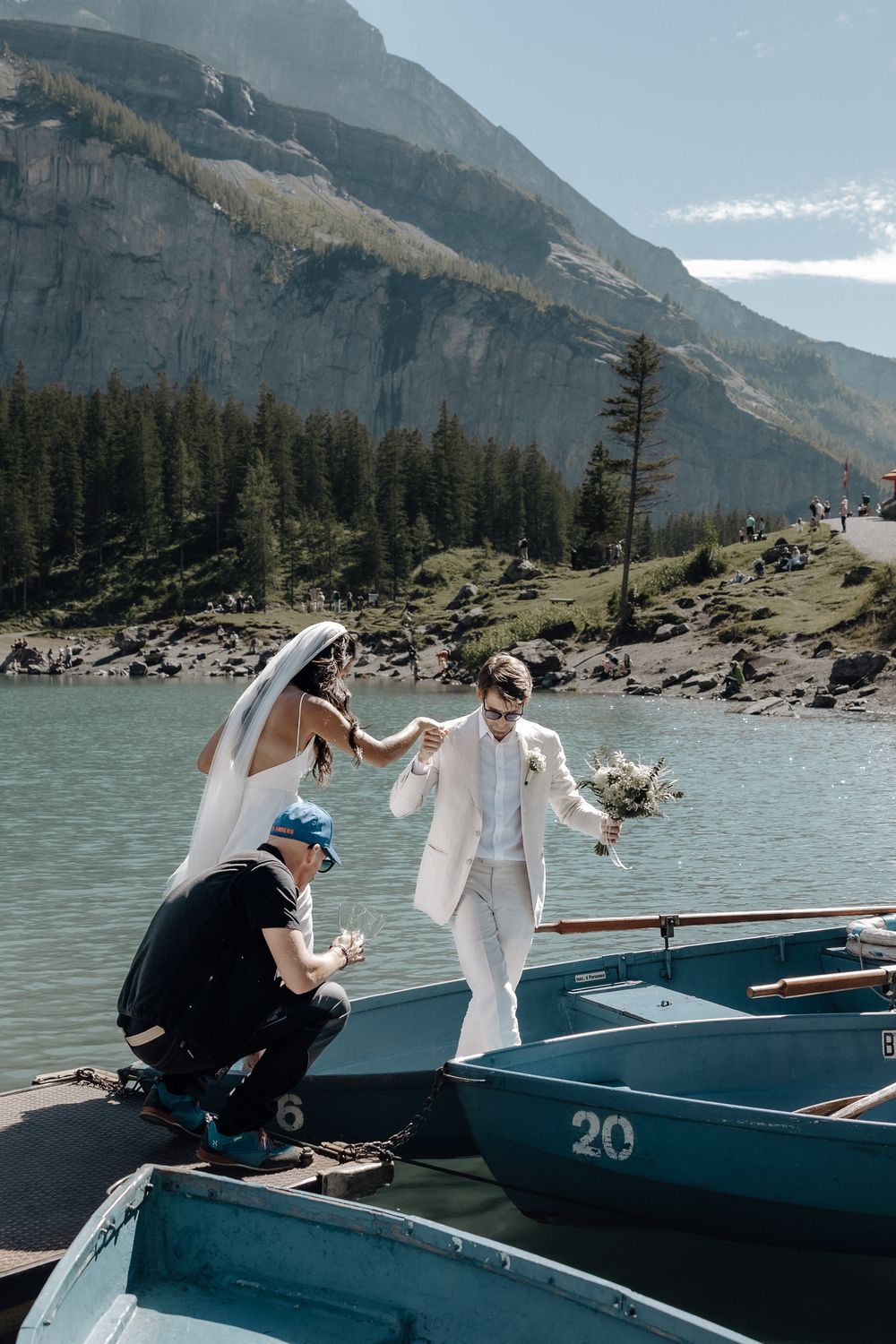 A couple boards a boat on a turquoise alpine lake surrounded by dramatic mountain cliffs and evergreen forest.