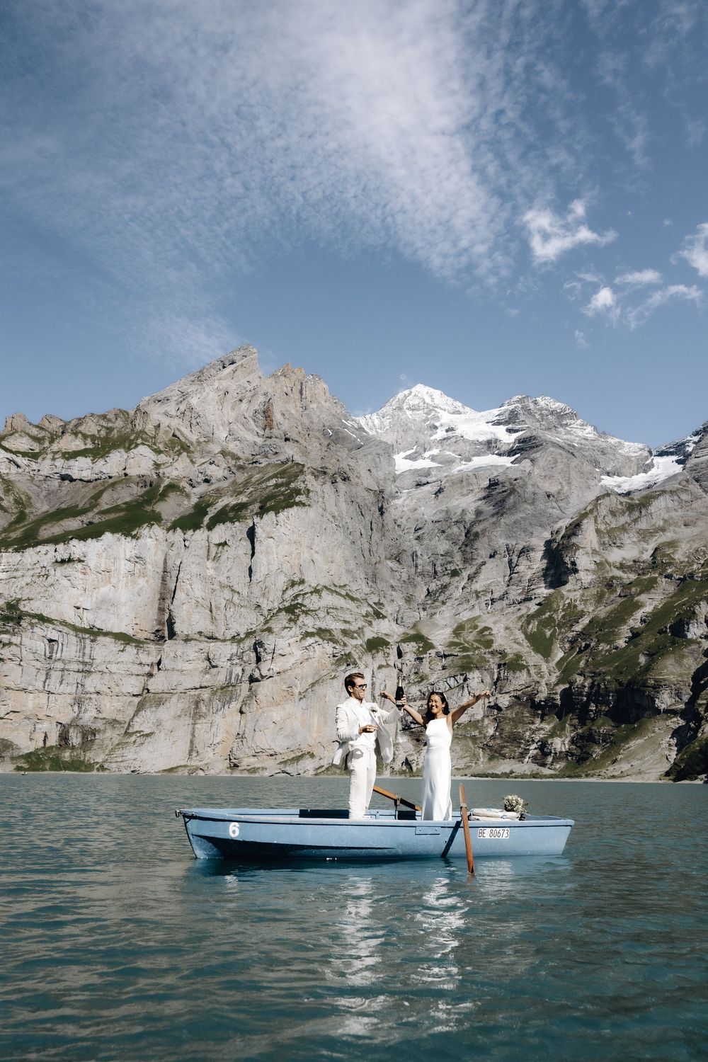 A romantic couple stands in a small blue boat on an alpine lake surrounded by dramatic snow-capped mountains.