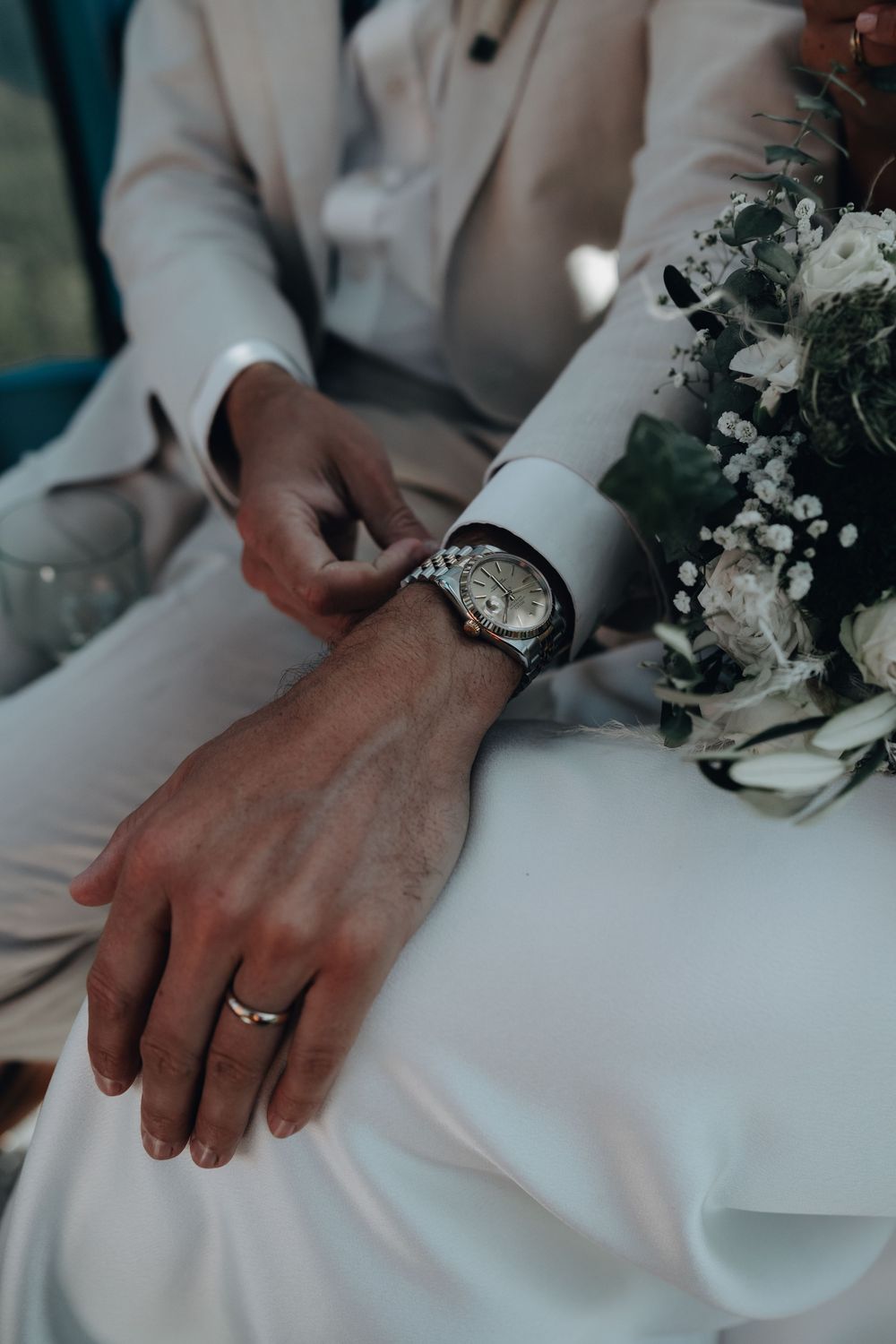 Wedding detail shot showing hands with rings and white floral arrangements on a table.