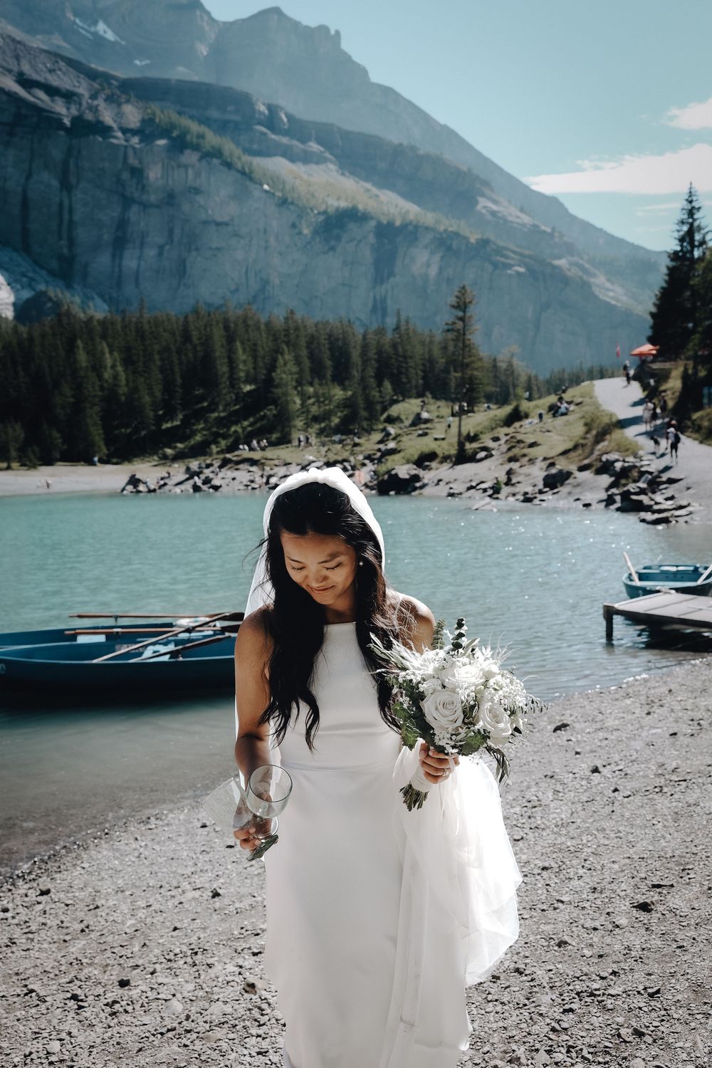 A bride in a white dress holds a bouquet on a rocky mountain lakeshore with snow-capped peaks and pine forests in the background.