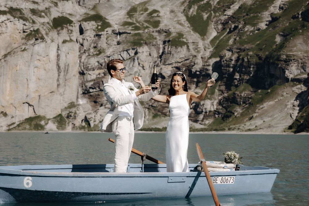 Couple wearing white outfits stands in blue boat on mountain lake during special celebration.