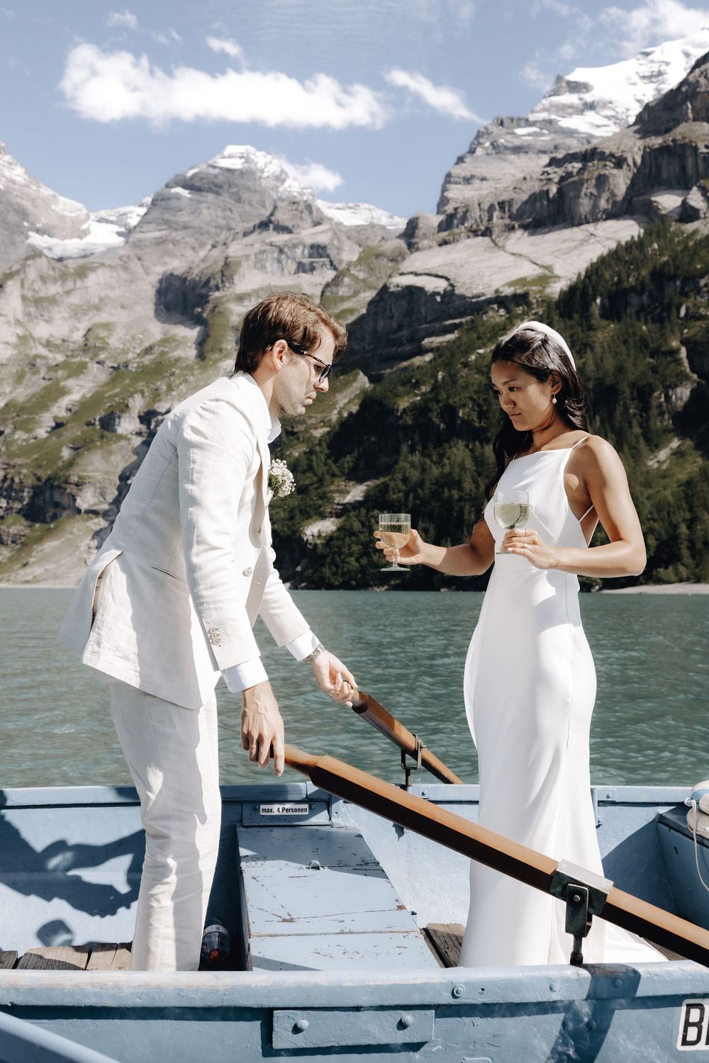 Couple in white outfits sharing a romantic moment on a boat with snowy mountains and lake in the background.