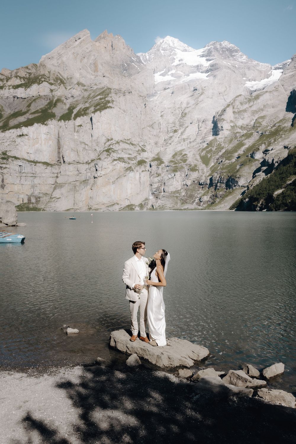 Intimate wedding moment captured on rocks by an alpine lake with snow-capped peaks in the distance.