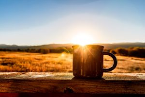 Steaming coffee cup silhouetted against golden sunset over autumn field with mountains in background.
