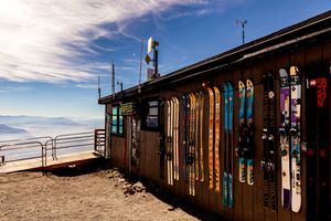 Ski and snowboard equipment lined up against a rustic mountain cabin with scenic snowy mountain views in the background.