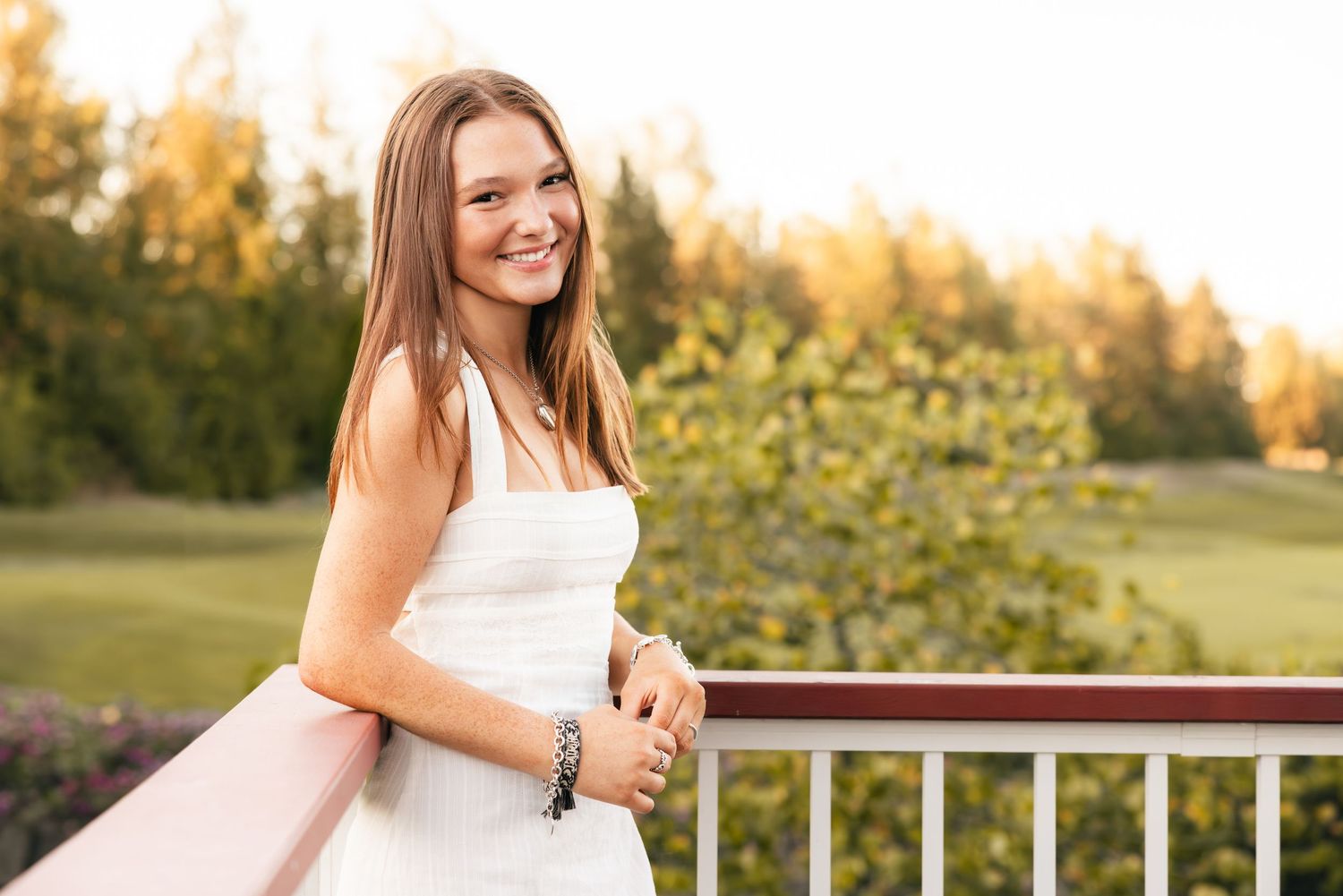 Someone in a white dress stands smiling on a deck railing during golden hour sunset in a garden setting.