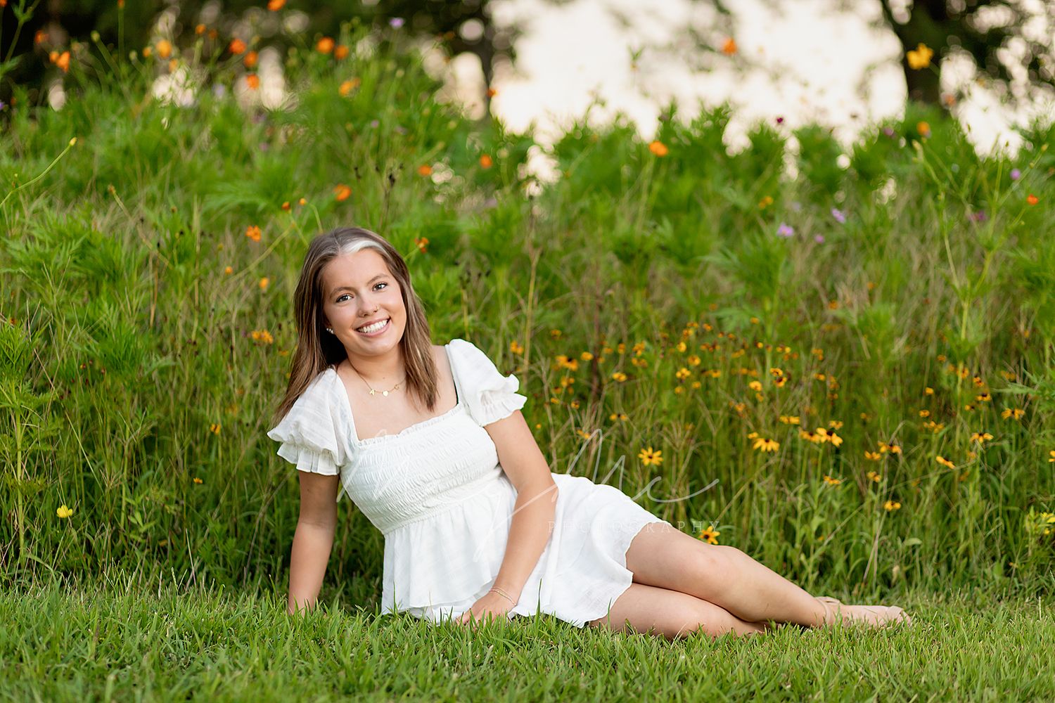 Teen in a white dress reclining on grass with wildflowers behind her on a summer day. Senior pictures with MegMc Photography in Medina, TN