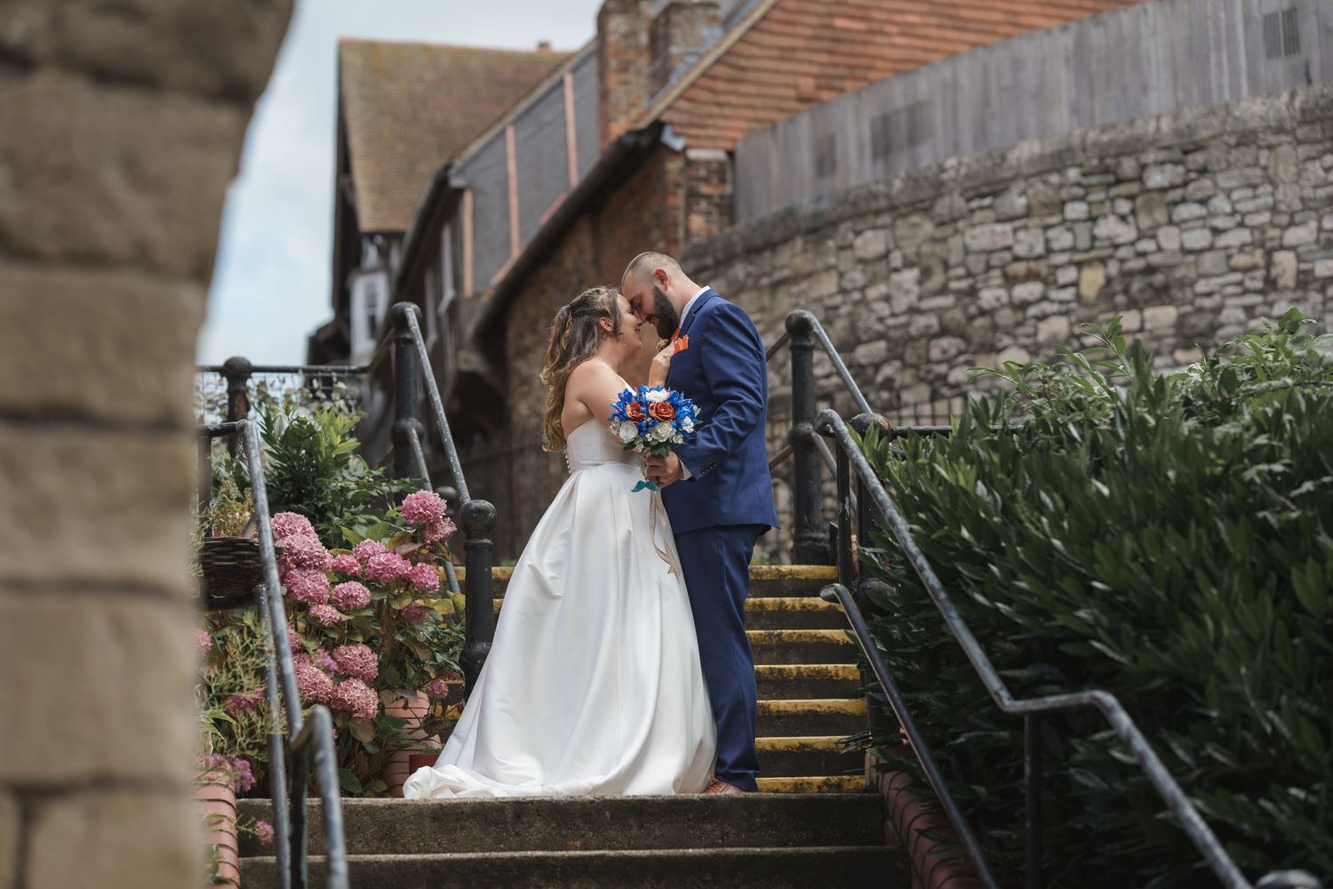 A couple shares a romantic moment on stone steps lined with greenery and pink flowers.