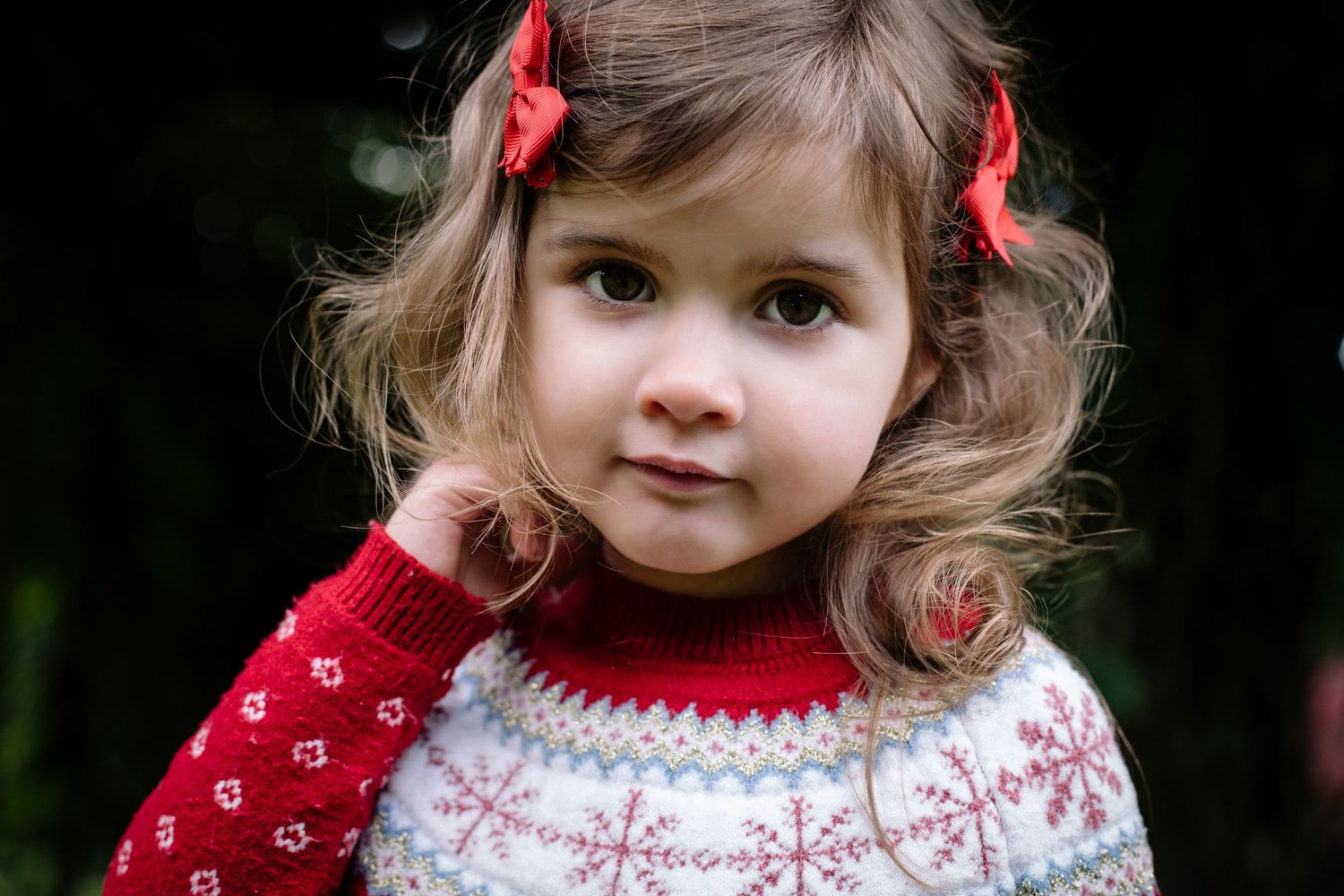 Young child wearing a festive red and white Nordic knit sweater with matching hair bows against dark background.