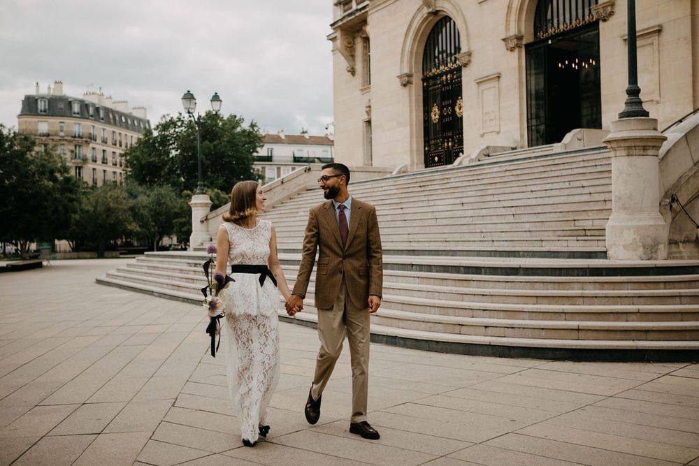Elegant newlywed couple walking hand in hand across a plaza in front of a grand Parisian building.