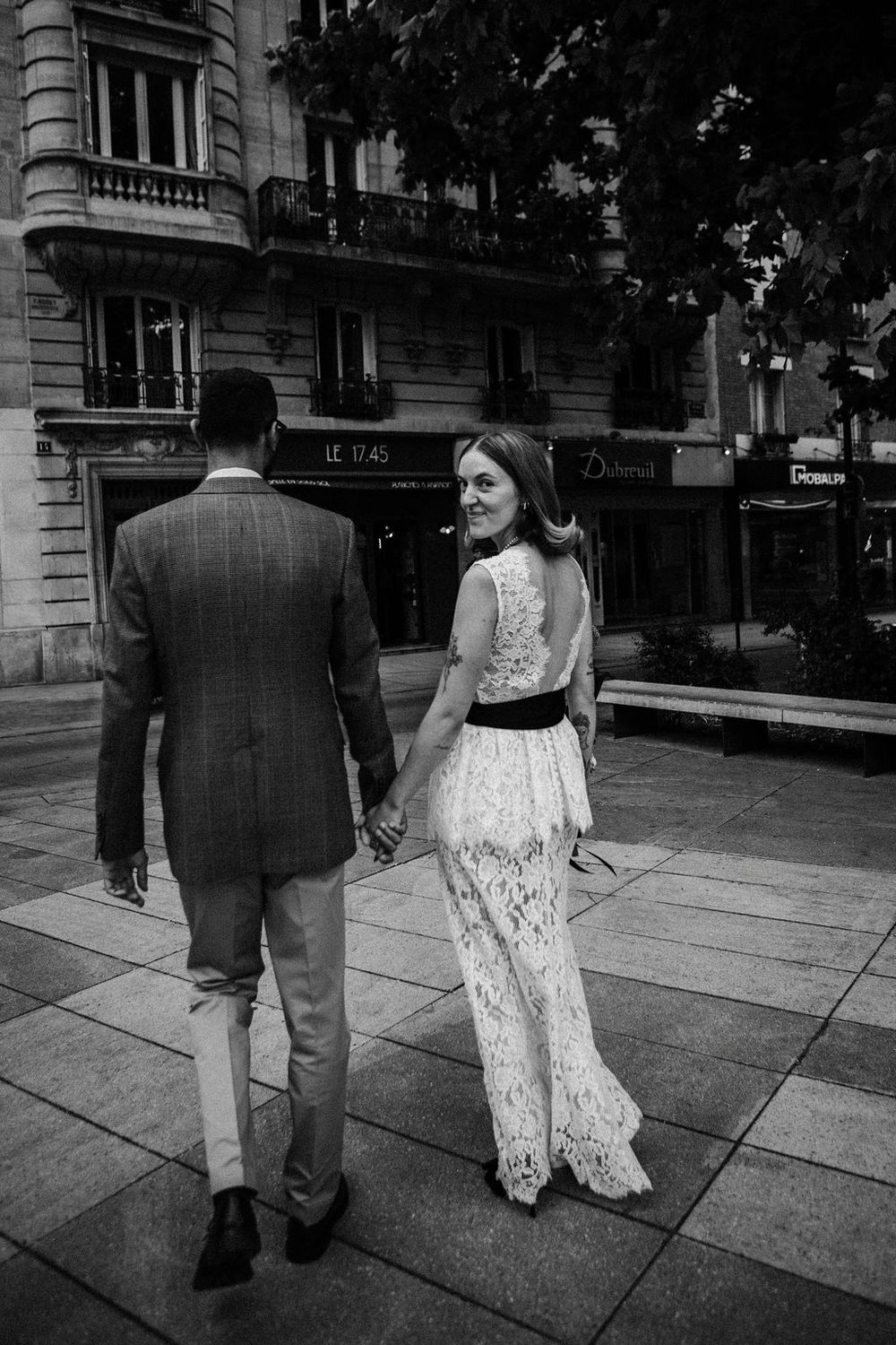 Black and white photo of bride in lace gown looking back while walking with groom on Paris street.