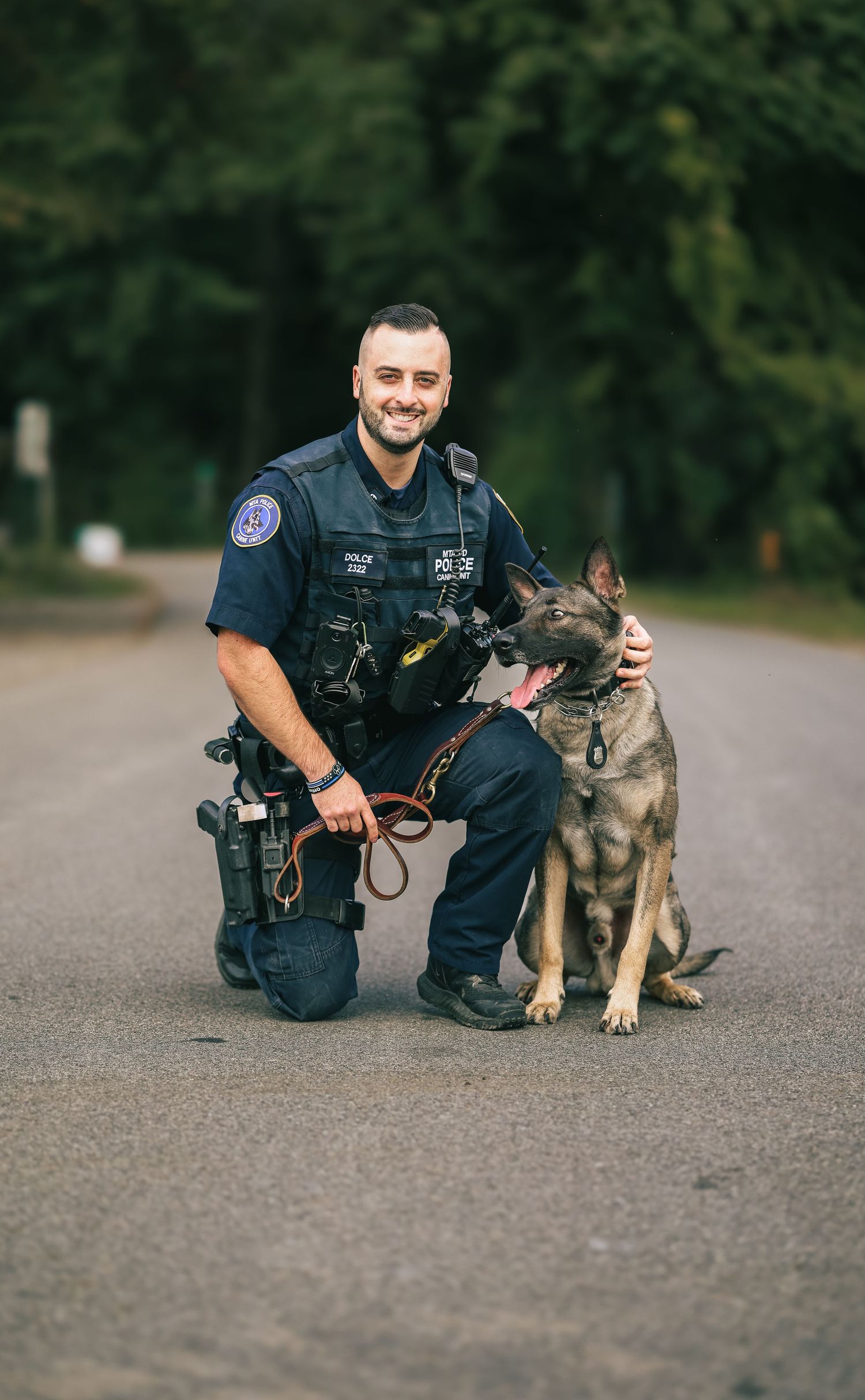 A police officer in uniform kneels beside a German Shepherd K9 partner on an asphalt surface during training.