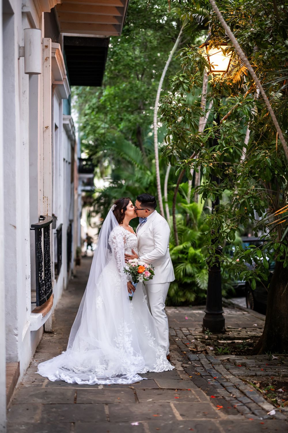 A romantic wedding couple embraces on a charming cobblestone street with a glowing lantern and lush greenery.
