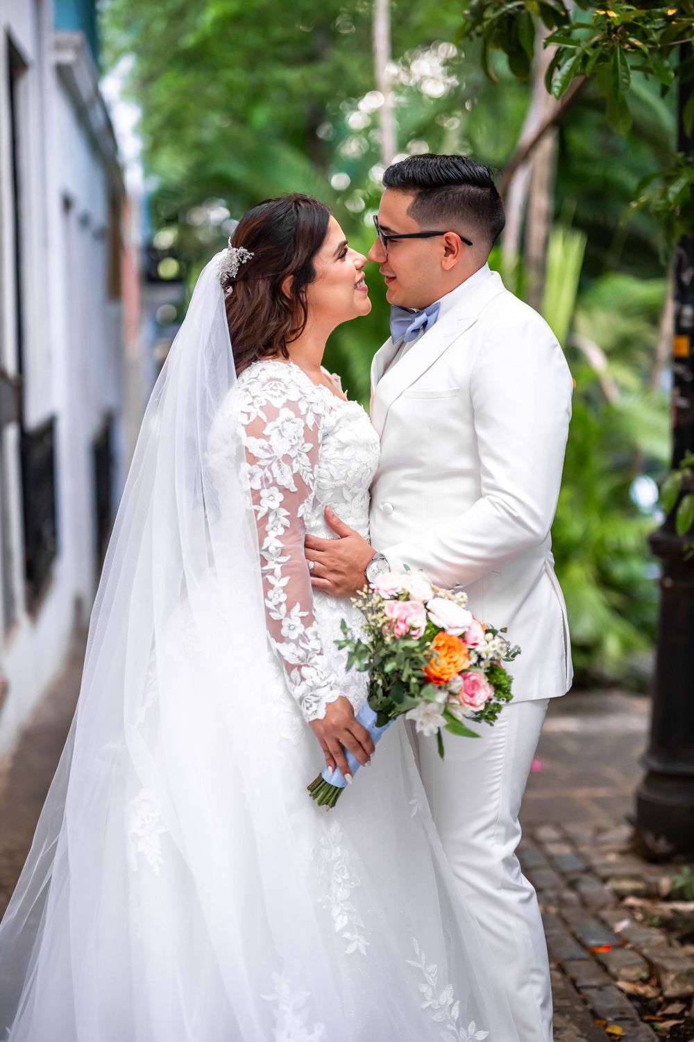 Wedding couple in white attire share romantic moments in an outdoor garden setting with beautiful floral bouquet.