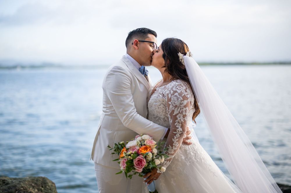 A couple in wedding attire share a romantic kiss by the water while holding a vibrant floral bouquet.