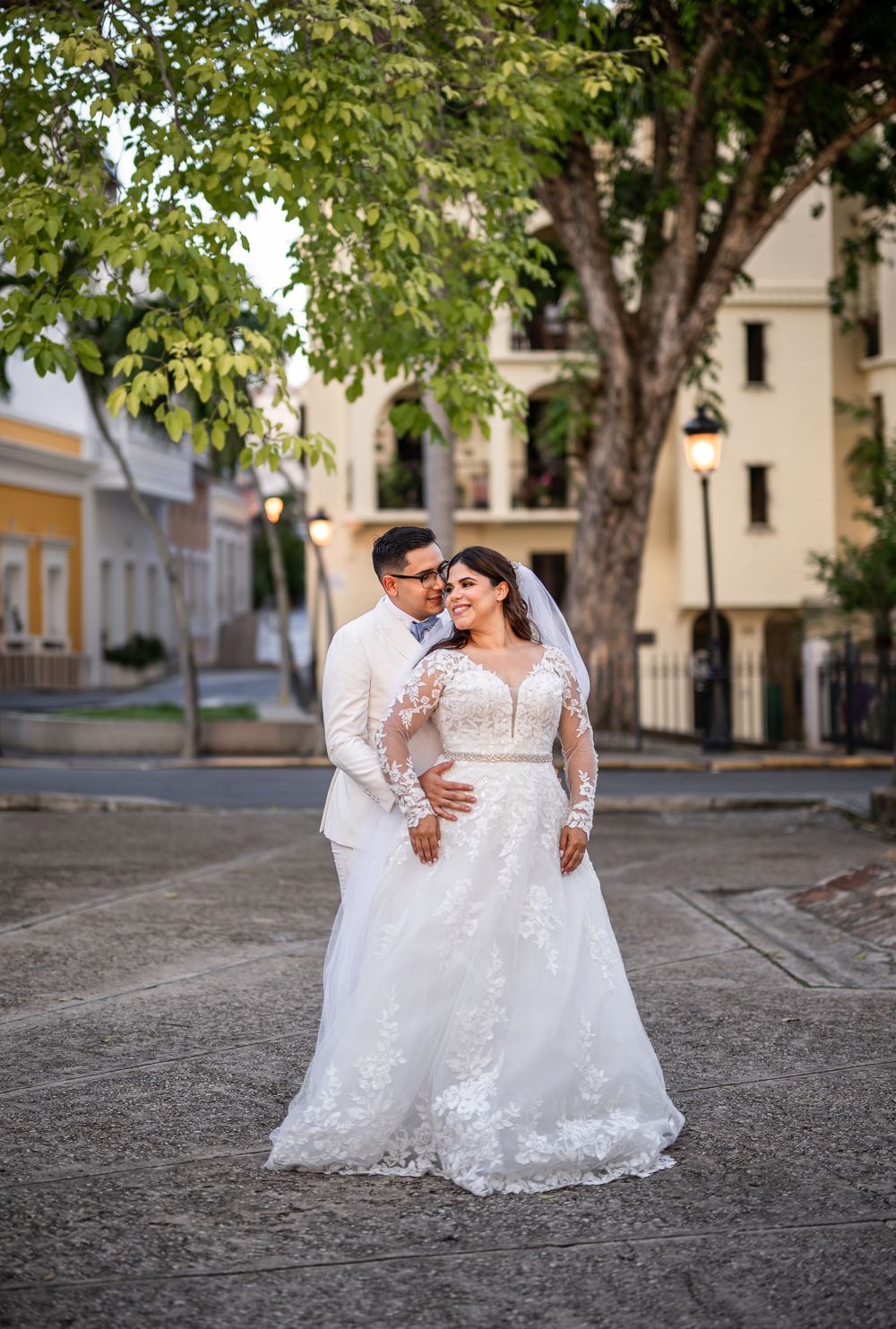A newlywed couple in white attire share a tender moment on a historic street with trees in the background.