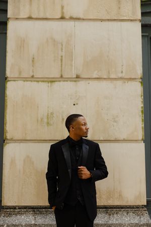 A figure in an elegant black suit and turtleneck poses against a weathered beige wall in a stylish portrait setting.