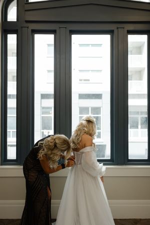 A tender moment captured by large windows as final adjustments are made to a flowing white wedding gown.