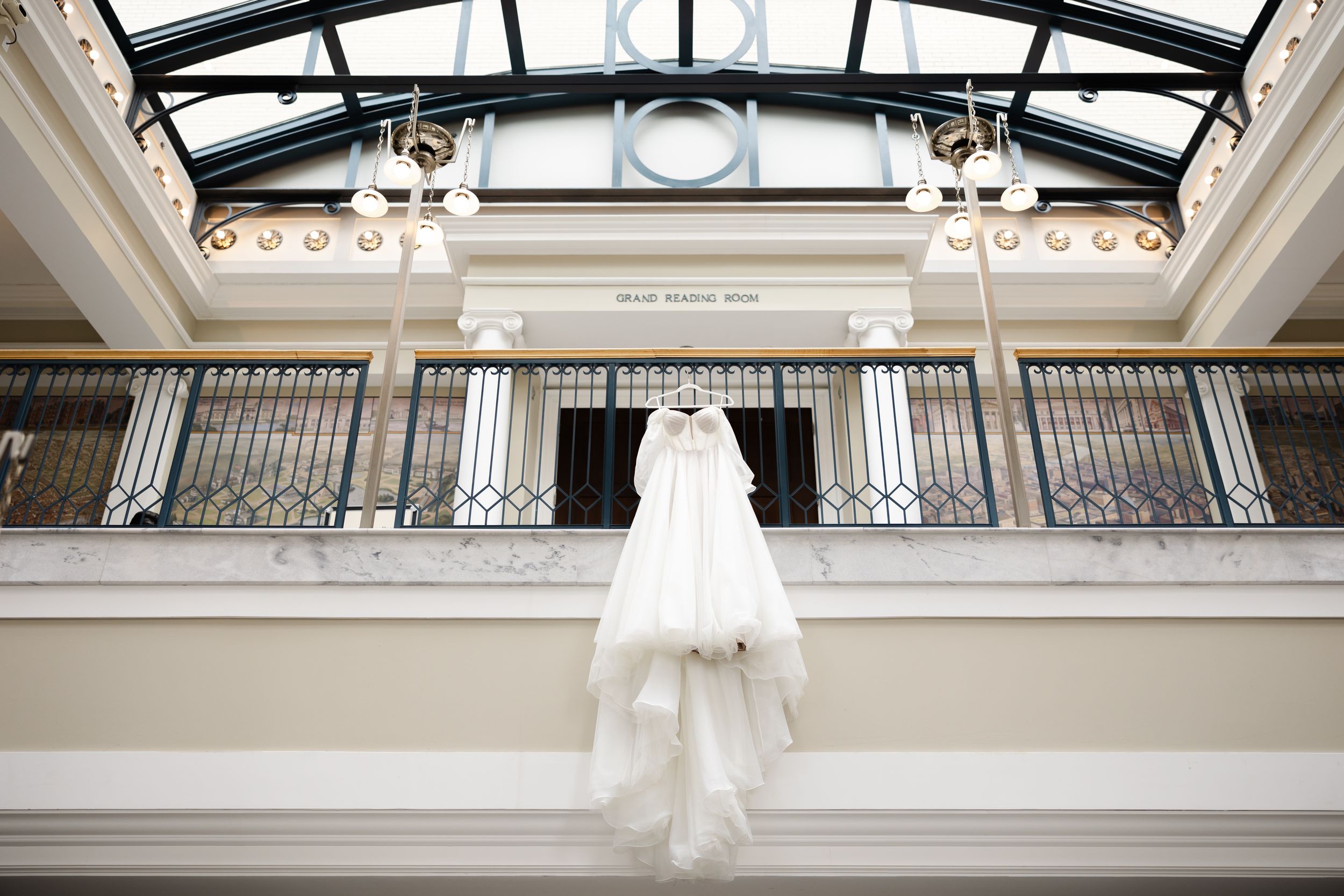 An elegant white wedding dress hangs from a balcony railing beneath a glass ceiling in a bright indoor venue.