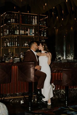 Newlyweds kissing at upscale bar, bride in white dress and groom in burgundy suit.